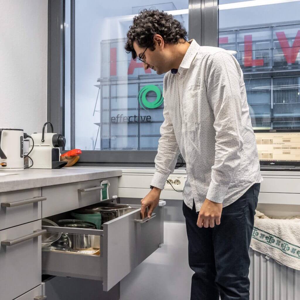 Ein Mann in weißem Hemd und schwarzer Hose steht in einer Küche und greift mit seiner rechten Hand nach einer offenen Schublade.